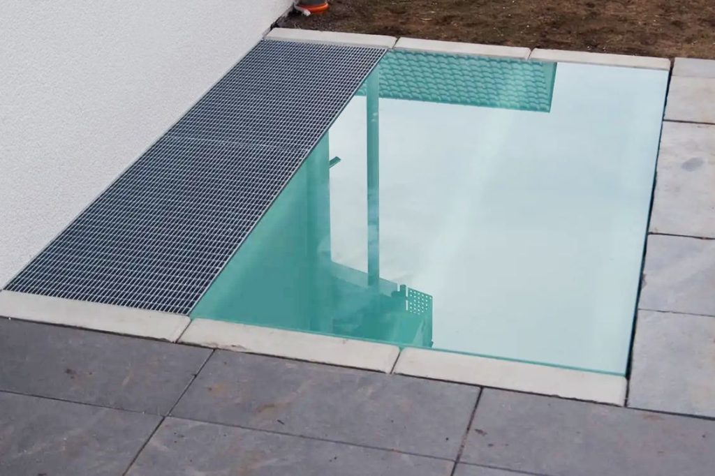 Begehbares Glas – modernes Design mit überraschend vielen Einsatzmöglichkeiten 1 Begehbares Glas als bodenbündige Lichtschachtabdeckung neben einem Gitterrost, eingefasst in Naturstein vor einer Hausfassade